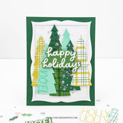 A handmade layered greeting card with green, yellow, and mint Christmas trees, silver glitter accents, and the words 'happy holidays' in white script. The card is displayed on a white surface with a few small decorative elements near the bottom.