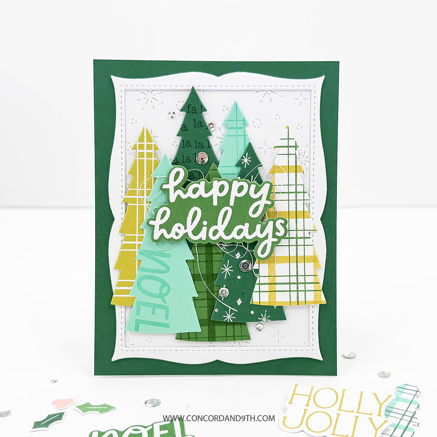 A handmade layered greeting card with green, yellow, and mint Christmas trees, silver glitter accents, and the words 'happy holidays' in white script. The card is displayed on a white surface with a few small decorative elements near the bottom.