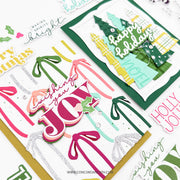 A close-up of layered Christmas greeting cards and decorative paper elements on a white background. The cards feature layered 3D letters spelling 'JOY', palm tree silhouettes, green Christmas trees, bows, holly, and phrases like 'wishing you a Merry Noel' and 'happy Holidays'.