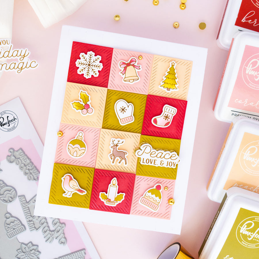A handmade Christmas greeting card displayed on a pink crafting surface, surrounded by ink pads, stamps, and small gold embellishments. The card features a 4x4 grid of holiday-themed square panels in red, pink, and olive green, with white die-cut elements including a snowflake, bells, a Christmas tree, holly, a mitten, a stocking, ornaments, a reindeer, a bird, a candle, and a cupcake.