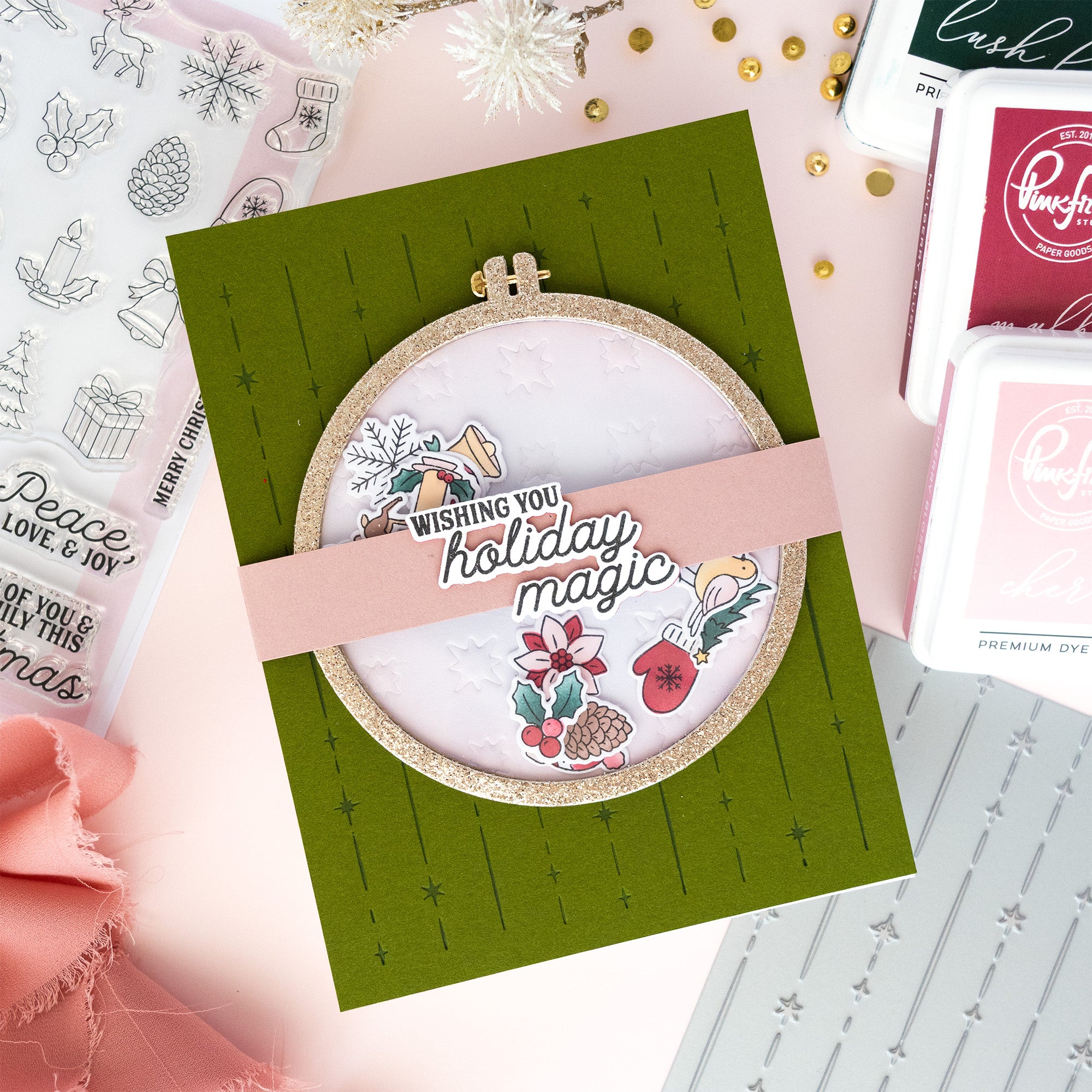 A handmade holiday greeting card featuring a circular embroidery hoop design with a pink banner reading 'Wishing you holiday magic,' surrounded by crafting tools and decorative elements on a pastel background.