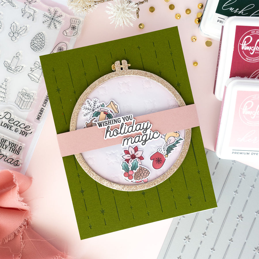 A handmade holiday greeting card featuring a circular embroidery hoop design with a pink banner reading 'Wishing you holiday magic,' surrounded by crafting tools and decorative elements on a pastel background.