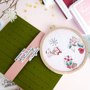A handmade holiday greeting card featuring a detachable circular embroidery hoop design with a pink banner reading 'Wishing you holiday magic,' surrounded by crafting tools and decorative elements on a pastel background.