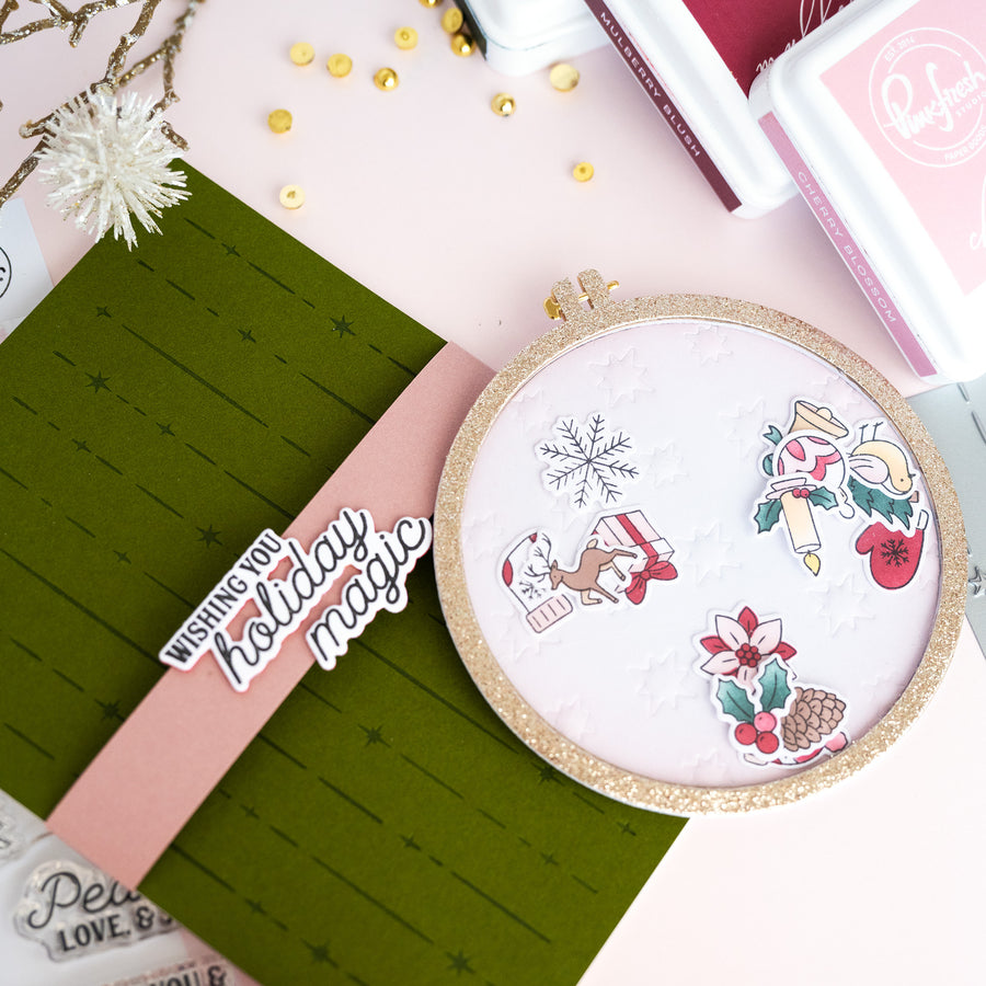 A handmade holiday greeting card featuring a detachable circular embroidery hoop design with a pink banner reading 'Wishing you holiday magic,' surrounded by crafting tools and decorative elements on a pastel background.