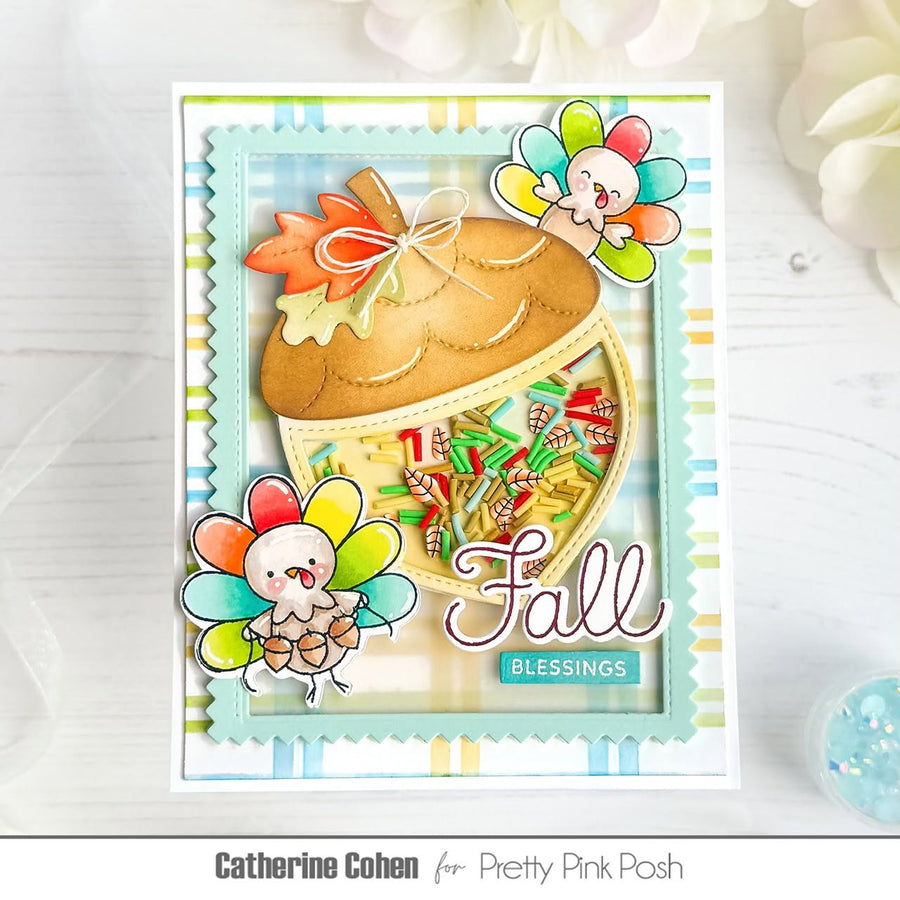 A handmade layered greeting card featuring a large acorn with a colorful confetti-filled window, autumn leaves, two cartoon turkeys, and the words 'Fall Blessings,' shown on a light wood surface with decorative flowers in the background.