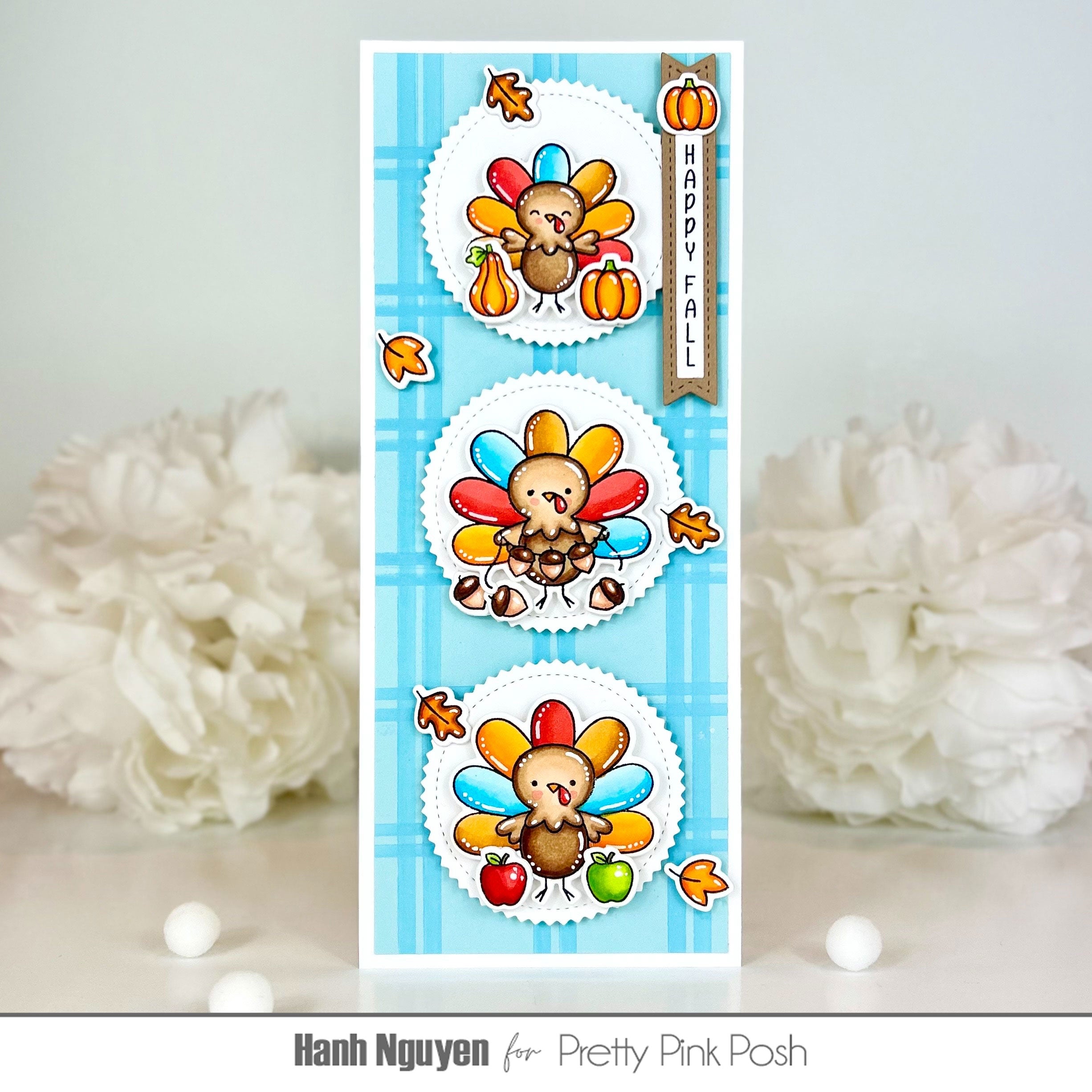 A handmade Thanksgiving-themed card featuring three cartoon turkeys with colorful feathers, pumpkins, autumn leaves, and a vertical banner reading 'HAPPY FALL' on a light blue plaid background. The card is photographed upright on a light surface with white decorative flowers blurred in the background.