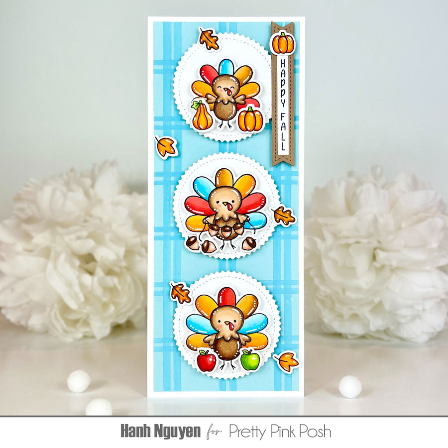 A handmade Thanksgiving-themed card featuring three cartoon turkeys with colorful feathers, pumpkins, autumn leaves, and a vertical banner reading 'HAPPY FALL' on a light blue plaid background. The card is photographed upright on a light surface with white decorative flowers blurred in the background.