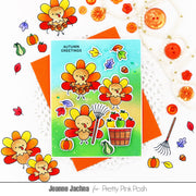 A flat-lay of a handmade autumn greeting card featuring cute cartoon turkeys, leaves, pumpkins, and a basket of fruit, styled with orange paper, lace, buttons, and fall decorations on a white background. The card reads 'AUTUMN GREETINGS' and is branded for Pretty Pink Posh by Jeanne Jachna.