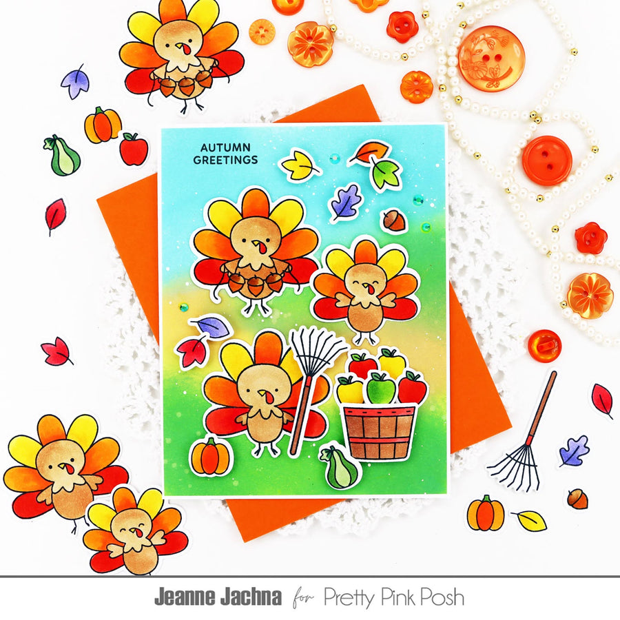 A flat-lay of a handmade autumn greeting card featuring cute cartoon turkeys, leaves, pumpkins, and a basket of fruit, styled with orange paper, lace, buttons, and fall decorations on a white background. The card reads 'AUTUMN GREETINGS' and is branded for Pretty Pink Posh by Jeanne Jachna.