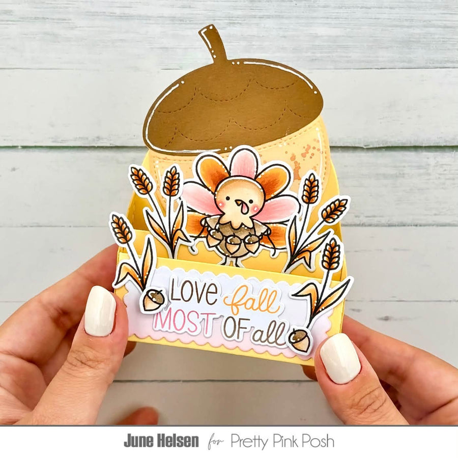 A handmade autumn-themed paper pop-up card held by two hands against a light gray wood plank background. The card features a die-cut acorn, a small cartoon bird, wheat stalks, and the text 'Love fall MOST OF all'.