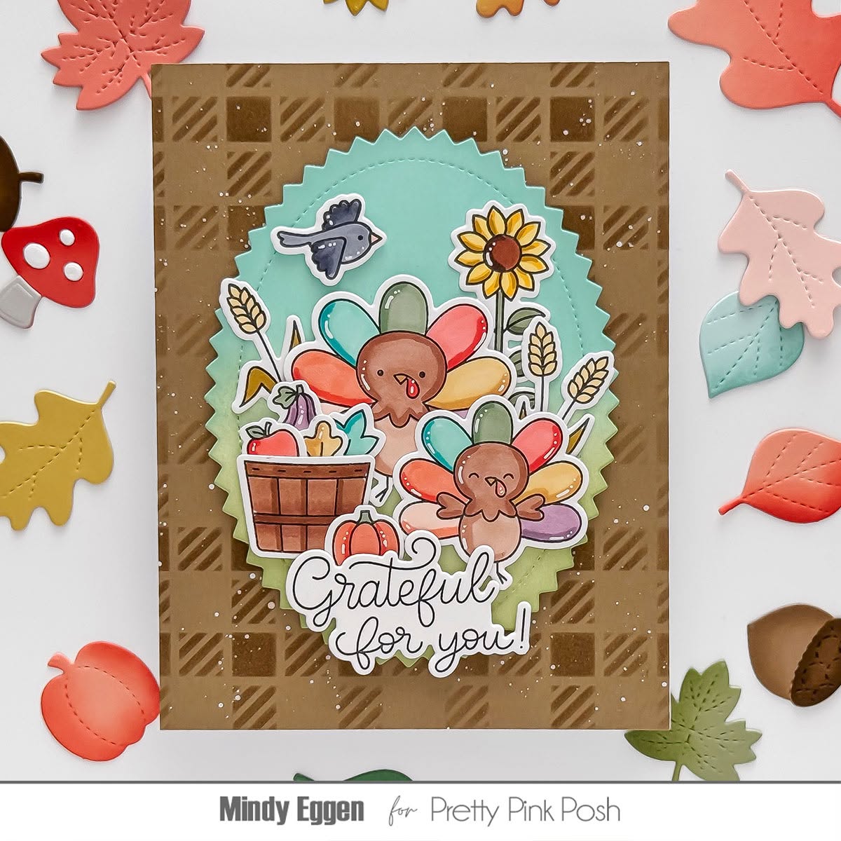 A handmade Thanksgiving-themed card featuring two colorful turkeys, a sunflower, acorns, and the phrase 'Grateful for you!' on a brown plaid background. The card is styled with autumn leaf and acorn cutouts around it, with the text 'Mindy Eggen for Pretty Pink Posh' at the bottom.