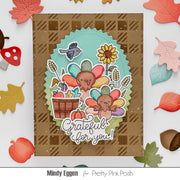 A handmade Thanksgiving-themed card featuring two colorful turkeys, a sunflower, acorns, and the phrase 'Grateful for you!' on a brown plaid background. The card is styled with autumn leaf and acorn cutouts around it, with the text 'Mindy Eggen for Pretty Pink Posh' at the bottom.