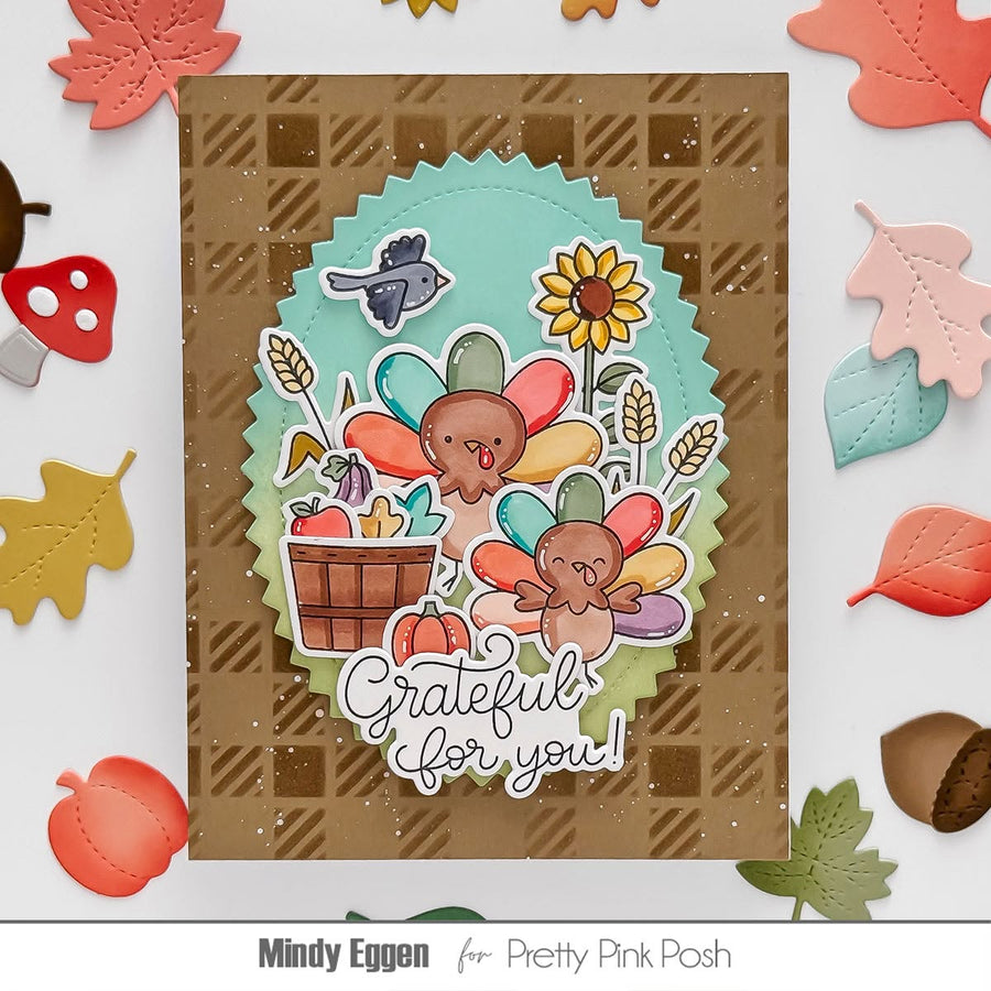 A handmade Thanksgiving-themed card featuring two colorful turkeys, a sunflower, acorns, and the phrase 'Grateful for you!' on a brown plaid background. The card is styled with autumn leaf and acorn cutouts around it, with the text 'Mindy Eggen for Pretty Pink Posh' at the bottom.