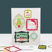 A handmade Christmas greeting card displayed on a white surface against a dark teal background. The card features layered die-cut frames with festive illustrations including a Christmas tree, Santa Claus, a snowflake, and a snowy house.