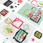 A styled flat-lay of Christmas-themed crafting supplies and cards on a white background, including layered holiday cards, scalloped die-cut frames, snowflake and star embellishments, and a small Santa card.