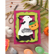 A handmade layered greeting card featuring a white dog dressed as a ghost with black bats holding an orange jack-o'-lantern. The card is layered over a green frame on a bright pink background and is displayed on a wooden surface with Halloween-themed props including scissors, cotton balls, leaves, and a small pumpkin.