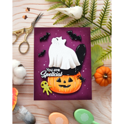 A handmade Halloween greeting card featuring a white ghost cat on top of a jack-o'-lantern, with the phrase 'You are Spellcial' in white script. The card is displayed in a flat-lay arrangement on a light wood background with autumn-themed props including a small orange pumpkin, green leaves, scissors, and cotton bolls.
