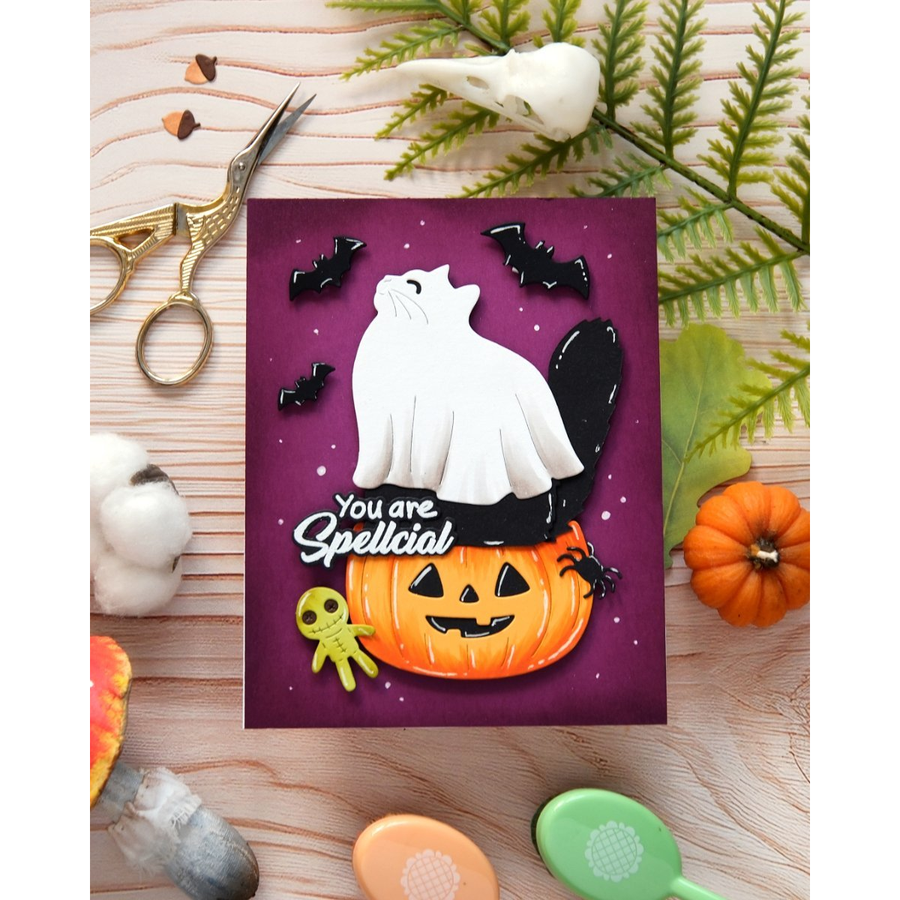 A handmade Halloween greeting card featuring a white ghost cat on top of a jack-o'-lantern, with the phrase 'You are Spellcial' in white script. The card is displayed in a flat-lay arrangement on a light wood background with autumn-themed props including a small orange pumpkin, green leaves, scissors, and cotton bolls.