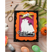 A handmade Halloween greeting card featuring a white cat dressed as a cat, an orange frame, and the text 'Witch-ing you a happy birthday' on a black background, styled on a wooden surface with autumn props.