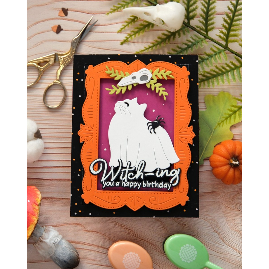 A handmade Halloween greeting card featuring a white cat dressed as a cat, an orange frame, and the text 'Witch-ing you a happy birthday' on a black background, styled on a wooden surface with autumn props.