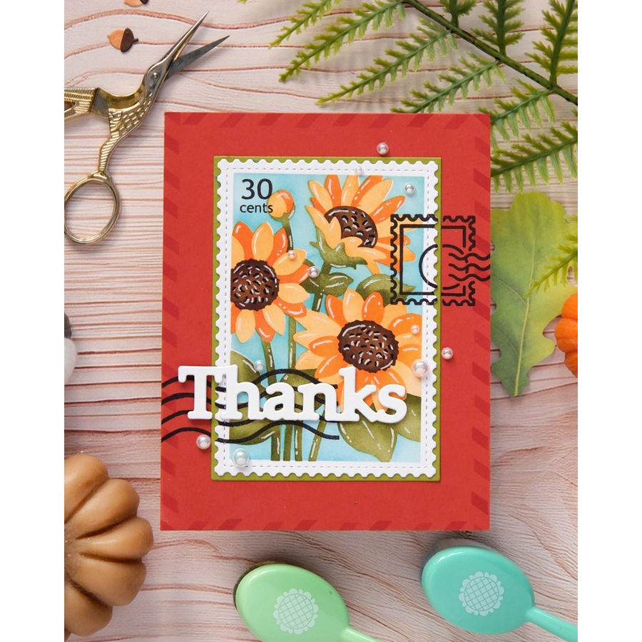 A handmade greeting card with a red border, a central sunflower illustration, a postage stamp motif, and the word 'Thanks' in white. The card is styled on a light wood surface with decorative props including scissors, leaves, and spoons.