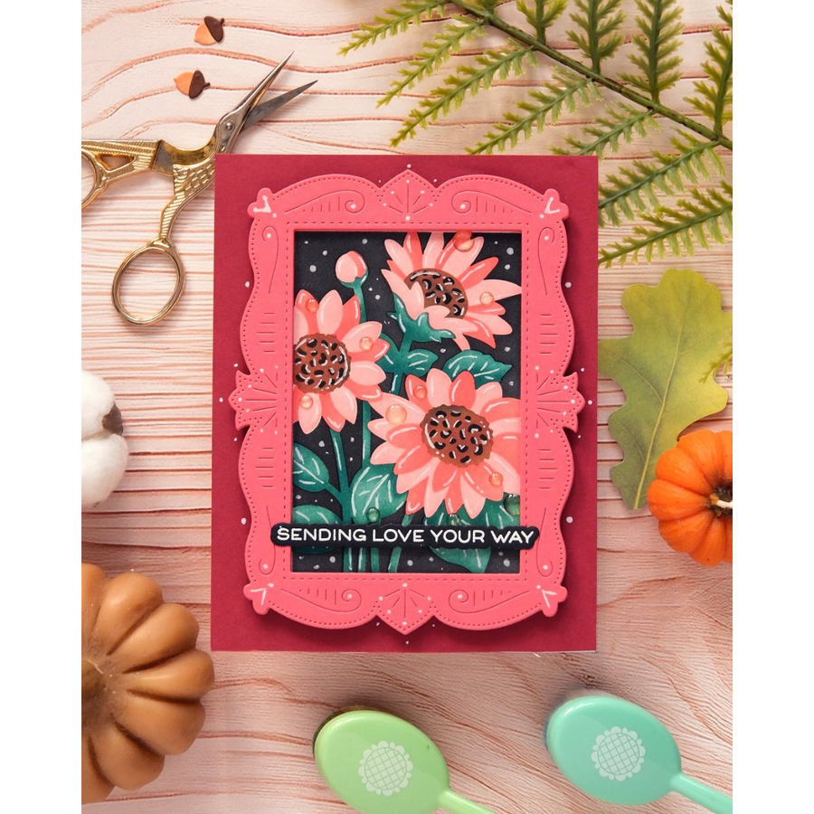 A handmade greeting card featuring layered pink scalloped frames and a central floral illustration of pink sunflowers on a dark background with the text 'SENDING LOVE YOUR WAY'. The card is styled in a flat-lay arrangement on a light wood surface with autumn-themed props including scissors, leaves, pumpkins, and spoons.