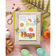 A handmade layered greeting card with the word 'Hugs' in 3D letters, decorated with white daisies, blue flowers, and orange pom-poms, styled on a light wood background with autumn props.