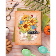 A handmade autumn-themed greeting card featuring layered paper sunflowers, a white grid basket, and a black banner that reads 'PROUD TO SEE YOU BLOOM'. The card is styled in a flat-lay arrangement with crafting scissors, greenery, a small pumpkin, and pastel paintbrushes on a light wood background.