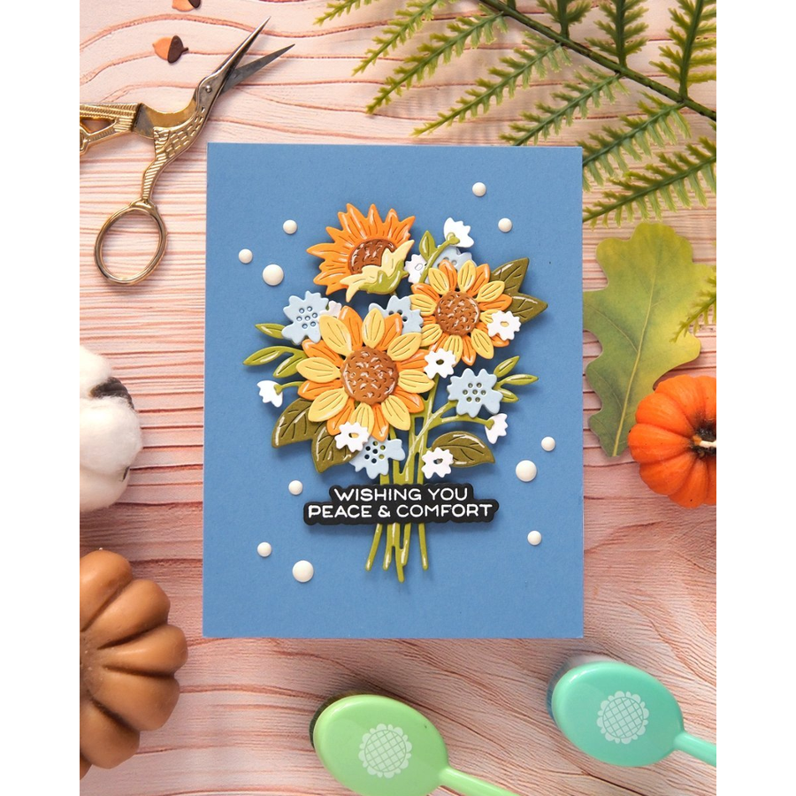 A layered paper craft card featuring a bouquet of yellow sunflowers, white blossoms, and green leaves on a dusty blue background, with the stamped text 'WISHING YOU PEACE & COMFORT'. The card is styled in a flat-lay arrangement on a light wood surface with autumn-themed props including a small pumpkin, scissors, and decorative spoons.