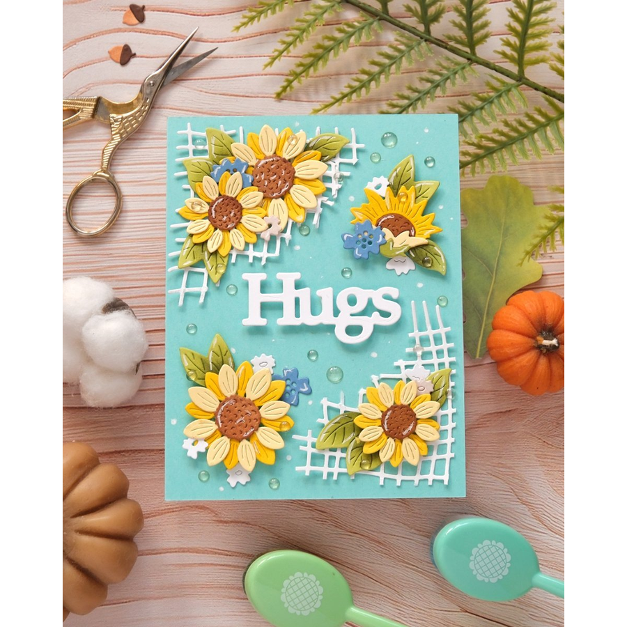 A handmade greeting card with a turquoise background, layered yellow and white sunflowers, white lattice details, and the word 'Hugs' in white. The card is styled in a flat-lay on a light wood surface with decorative props including scissors, cotton, leaves, and small pumpkins.