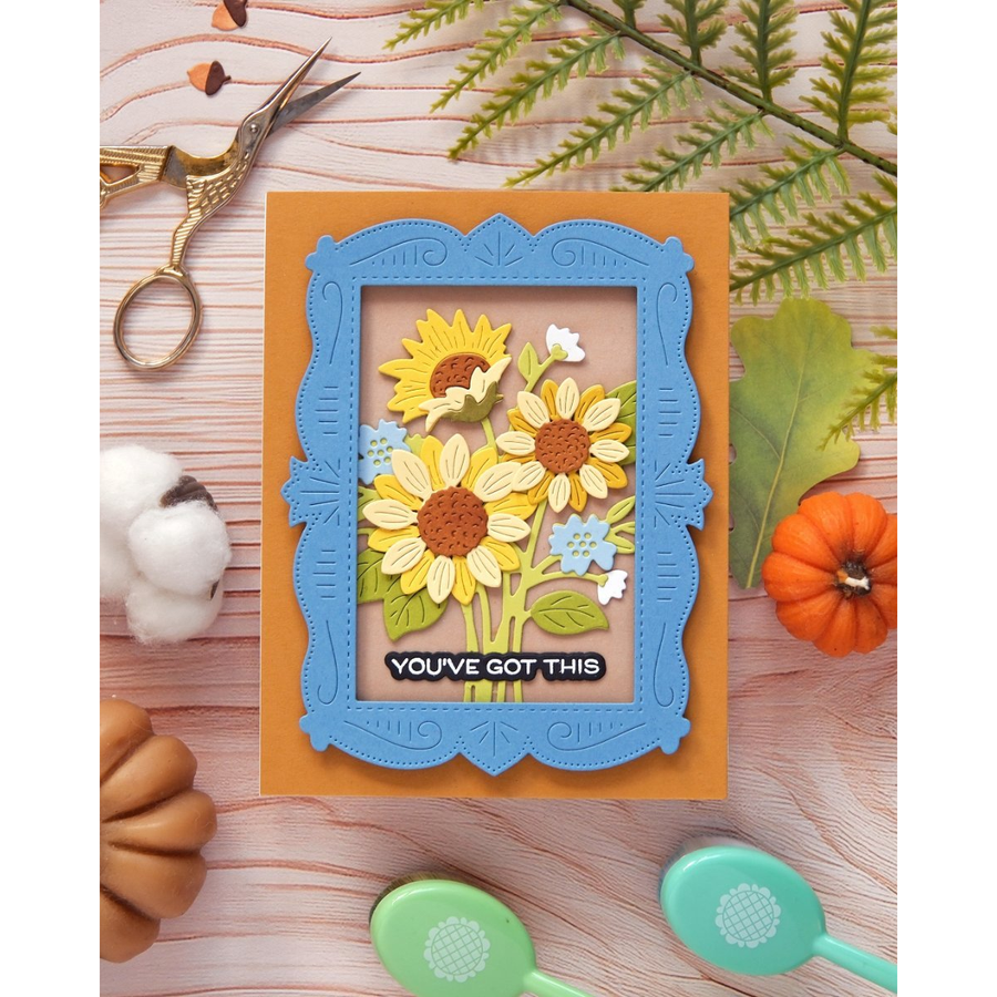 A handmade layered card featuring yellow and white sunflowers with green stems, set against a light pink background with a blue ornate frame. The card is placed on an orange backing and styled with autumn-themed props including a small pumpkin, cotton bolls, green leaves, scissors, and paintbrushes on a light wood surface.