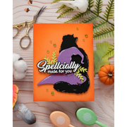 An orange rectangular card or plaque featuring a black cat silhouette sitting on a purple witch hat, with the phrase 'Spellcially made for you' in white script. The item is styled on a light wood surface with autumn props including a small pumpkin, leaves, scissors, cotton balls, and pastel spoons.
