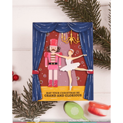 A Christmas-themed handmade greeting card featuring a nutcracker and ballerina under blue curtains, styled with pine branches and small red ornaments on a white wood background.