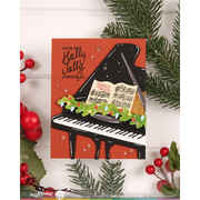 A handmade Christmas greeting card featuring a black grand piano with an open sheet music book, decorated with a green garland and red berries, set against a rust-orange background. The card is styled on a white wood background with pine branches and red berries.