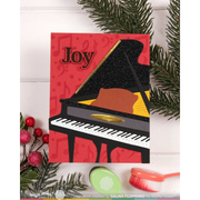 A Christmas-themed handmade greeting card featuring a black glittery grand piano with the word 'Joy' on a red background, styled with green pine branches and holiday decorations on a white wood panel backdrop.