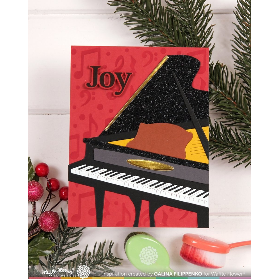 A Christmas-themed handmade greeting card featuring a black glittery grand piano with the word 'Joy' on a red background, styled with green pine branches and holiday decorations on a white wood panel backdrop.