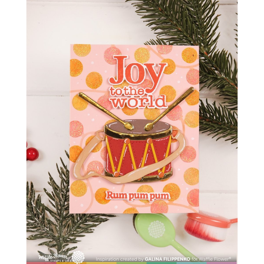 A festive handmade greeting card featuring a red toy drum with gold accents and the text 'Joy to the world' and 'Rum pum pum'. The card is styled with green pine branches, a small red ornament, and a green paintbrush on a white wood background.