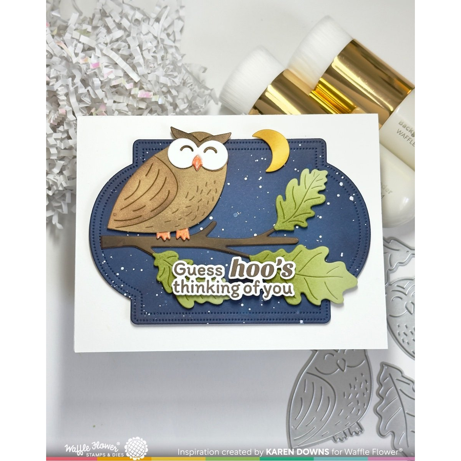 A handmade greeting card featuring a die-cut owl, moon, and oak leaves on a navy blue night-sky shape, with the phrase 'Guess hoo's thinking of you' on a white card. The card is styled with craft tools and paper shred in the background.