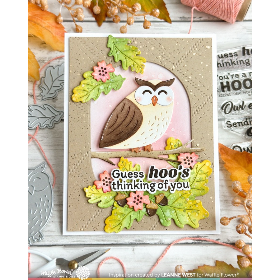 A handmade layered card featuring a brown and cream owl, pink flowers, and autumn leaves, with the text 'Guess hoo's thinking of you'. The card is displayed on a rustic white wood background with crafting props.