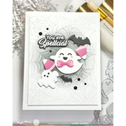 A handmade Halloween greeting card featuring a white ghost with a pink bow tie, a smaller ghost, bats, spiderwebs, and the text 'You are Spellcial'. The card is displayed on a light surface with craft props and metallic accents.