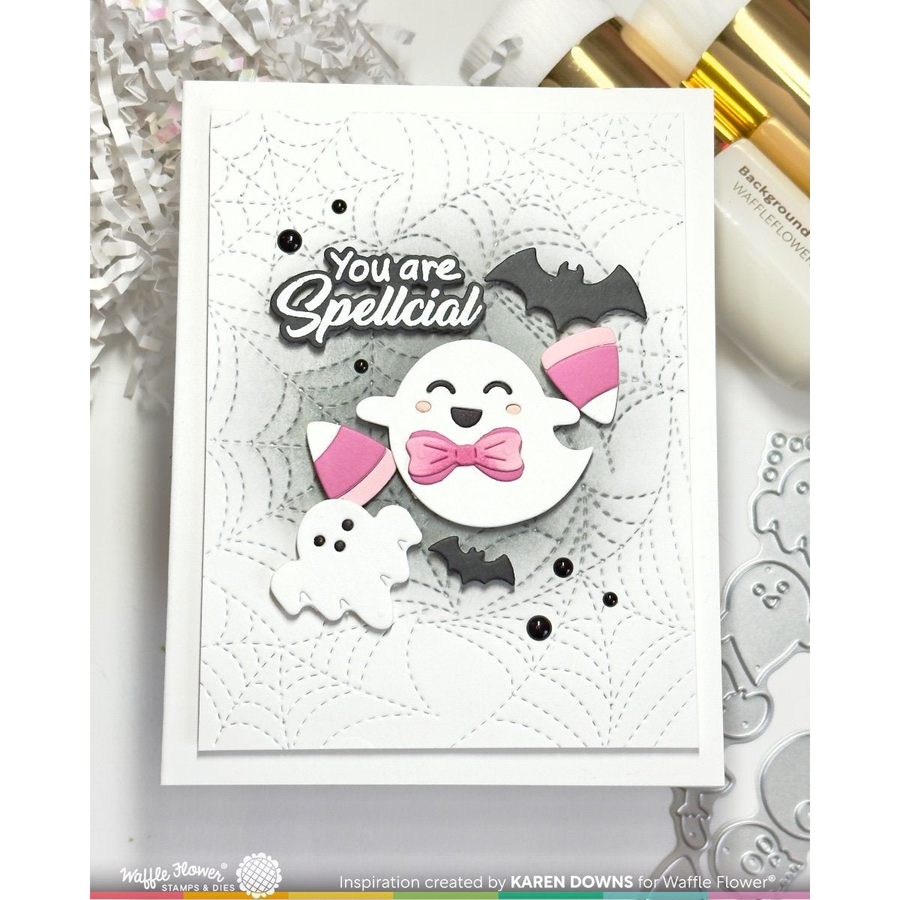 A handmade Halloween greeting card featuring a white ghost with a pink bow tie, a smaller ghost, bats, spiderwebs, and the text 'You are Spellcial'. The card is displayed on a light surface with craft props and metallic accents.
