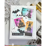 A handmade Halloween-themed card featuring layered die-cut stickers of haunted houses, ghosts, bats, and a skull on a white stamped background with black ink splatters. The card is displayed on a light gray surface with craft tools and paper shimmers in the background.