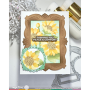 A handmade greeting card featuring stencilled sunflowers and decorative layered frames, styled on a white surface with crafting tools and paper around it. The card reads 'Wishing You Peace & Comfort' and shows Waffle Flower branding.