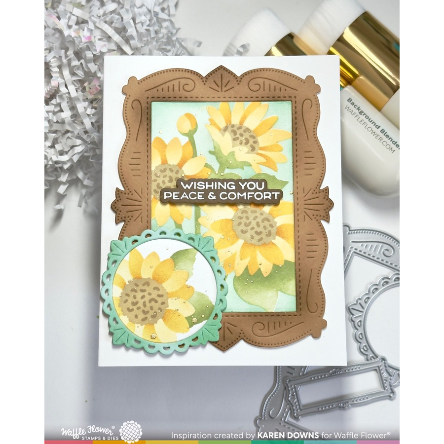 A handmade greeting card featuring stencilled sunflowers and decorative layered frames, styled on a white surface with crafting tools and paper around it. The card reads 'Wishing You Peace & Comfort' and shows Waffle Flower branding.