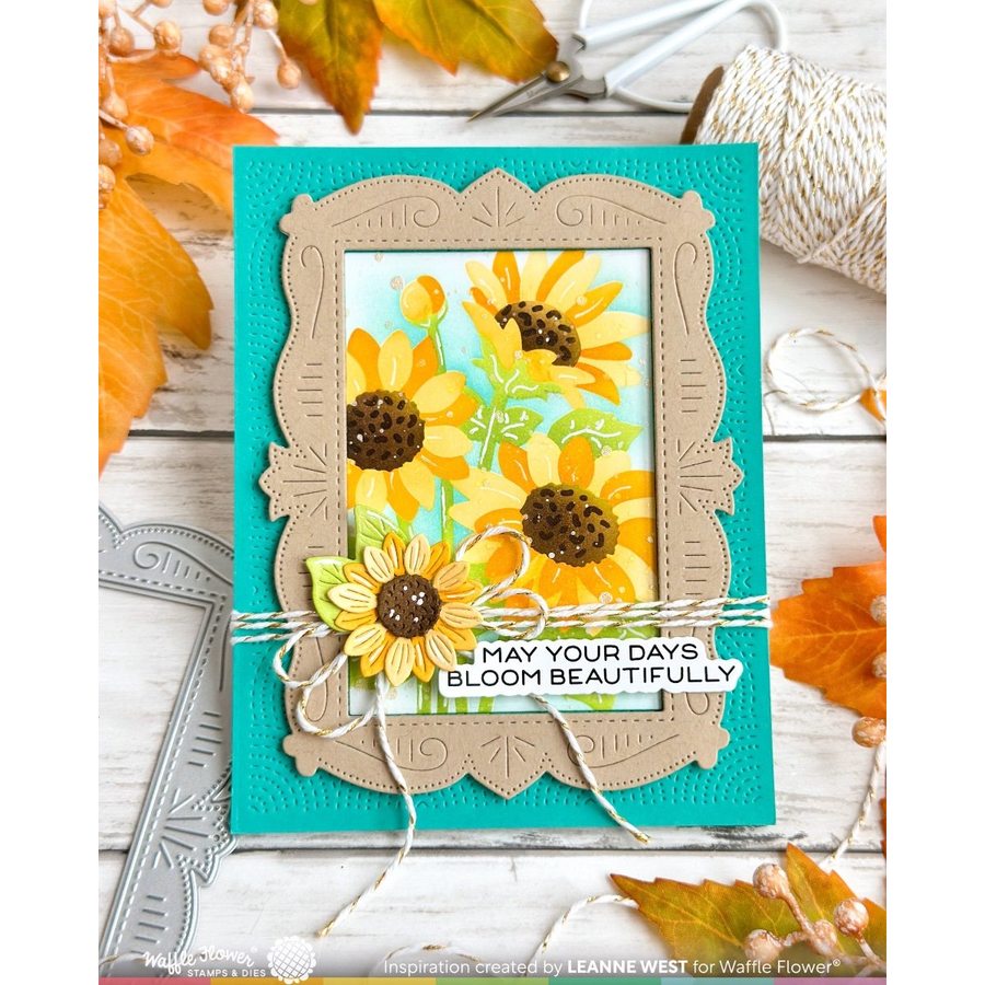 A handmade greeting card featuring a turquoise background, a beige decorative frame, and a layered sunflower design with a 3D flower and twine bow. The card is displayed on a white wood background with autumn leaves, scissors, and spools of thread.