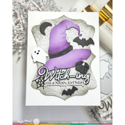 A handmade Halloween-themed greeting card featuring a purple witch hat, black bats, a small white ghost, and a pumpkin on a scalloped white background with the text 'Witching you a happy birthday'. The card is displayed on a light surface with craft supplies nearby.