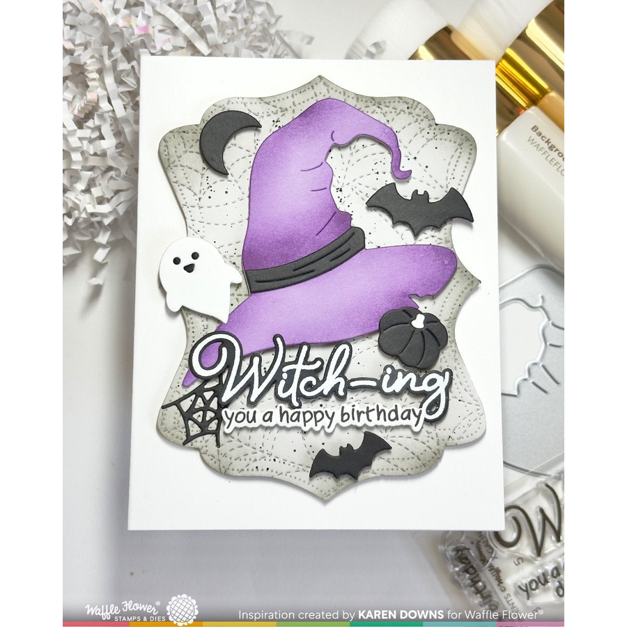 A handmade Halloween-themed greeting card featuring a purple witch hat, black bats, a small white ghost, and a pumpkin on a scalloped white background with the text 'Witching you a happy birthday'. The card is displayed on a light surface with craft supplies nearby.