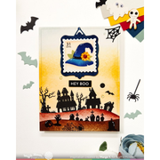 A Halloween-themed layered card featuring a blue witch hat stamp with a yellow flower, black haunted houses, tombstones, bats, and spiders over a yellow-to-orange gradient background. The card is styled with spooky paper cutouts and props on a white surface.