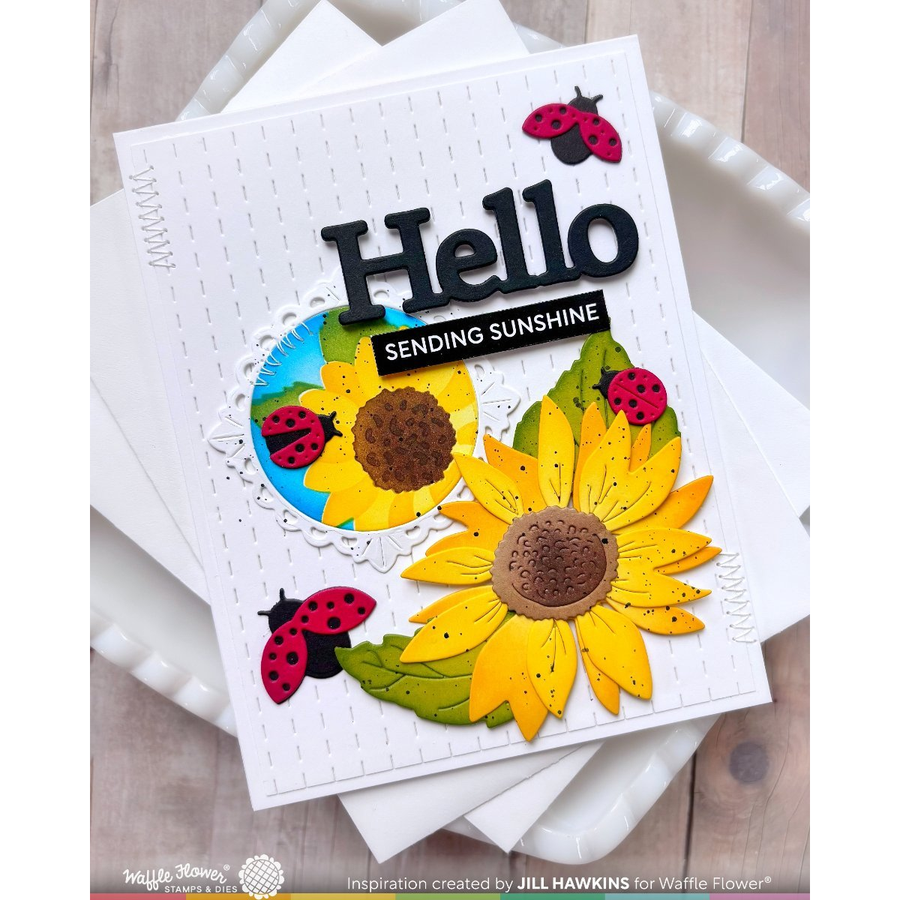 A handmade greeting card featuring yellow sunflowers, green leaves, ladybugs, and the text 'Hello Sending Sunshine' on a white card, styled on top of white envelopes over a light wood surface.