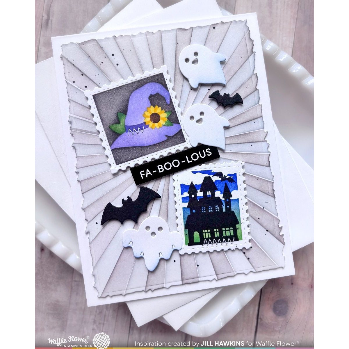 A handmade Halloween greeting card with layered white and gray sunburst panels, ghost cutouts, black bats, a purple witch hat with a yellow flower, a black haunted house stamp-style design, and a black banner reading 'FA-BOO-LOUS,' photographed on stacked white envelopes over a wood surface.